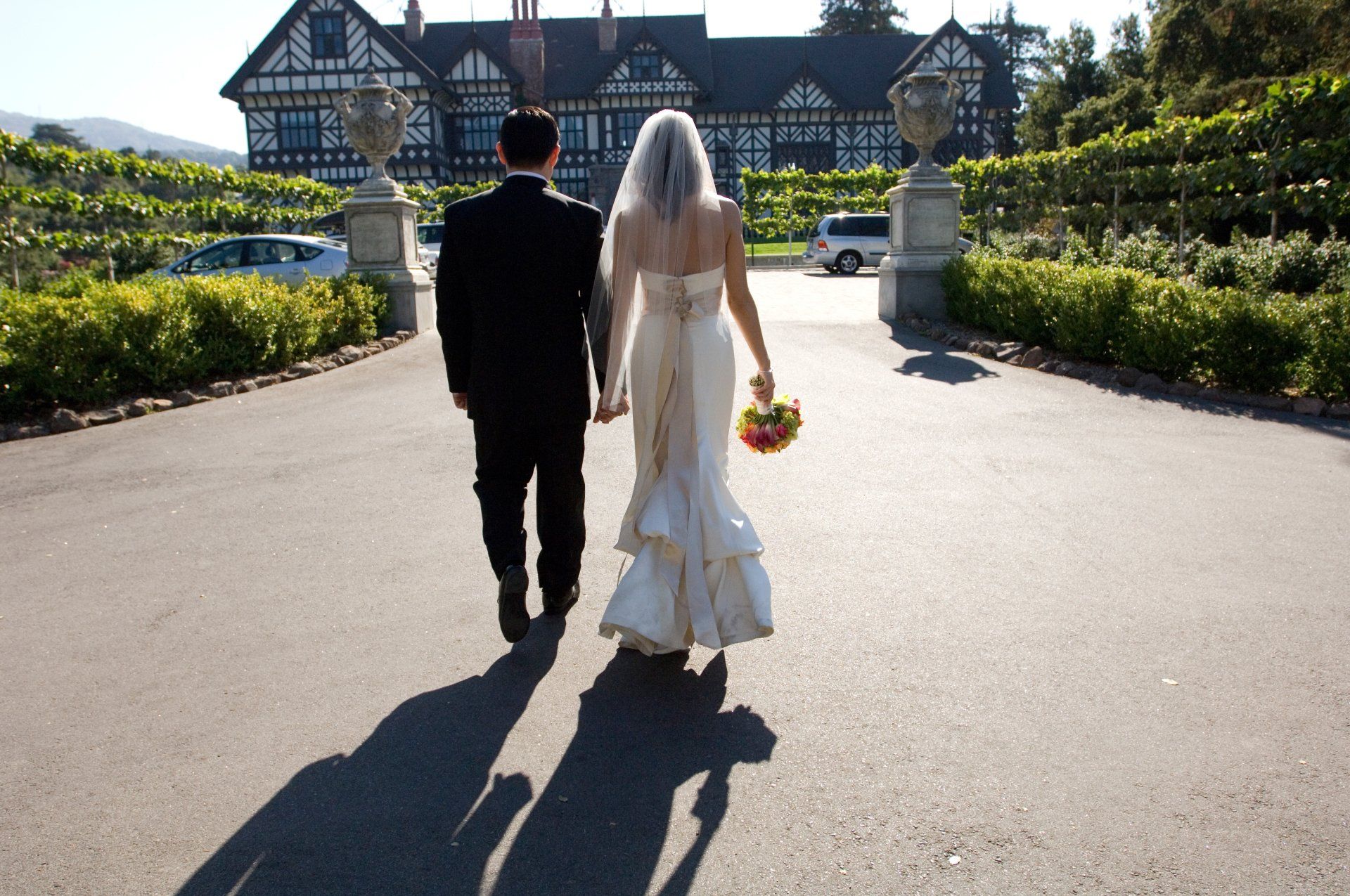 A bride and groom are walking down a road in front of a large house