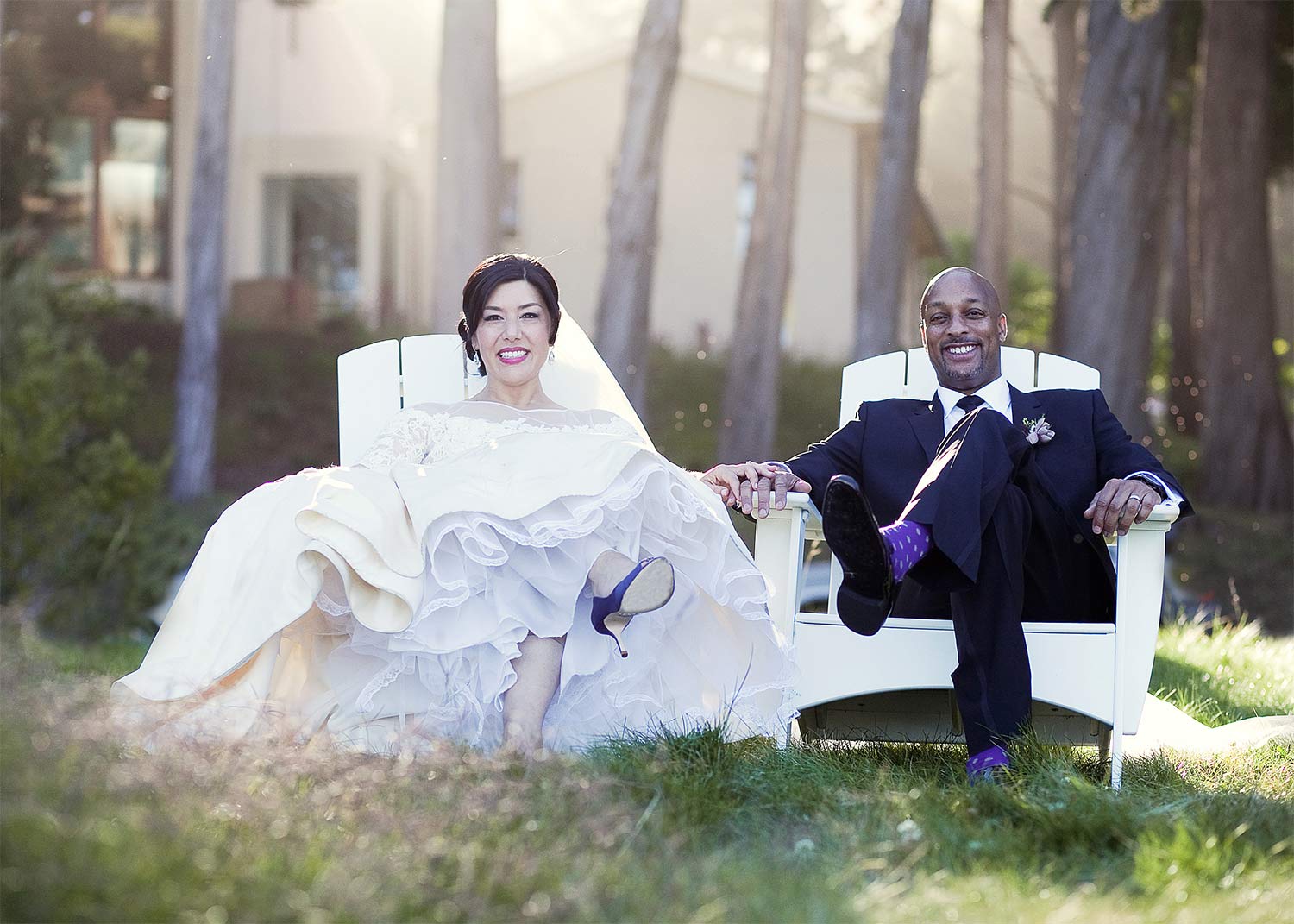 A bride and groom are sitting in white chairs in the grass holding hands.