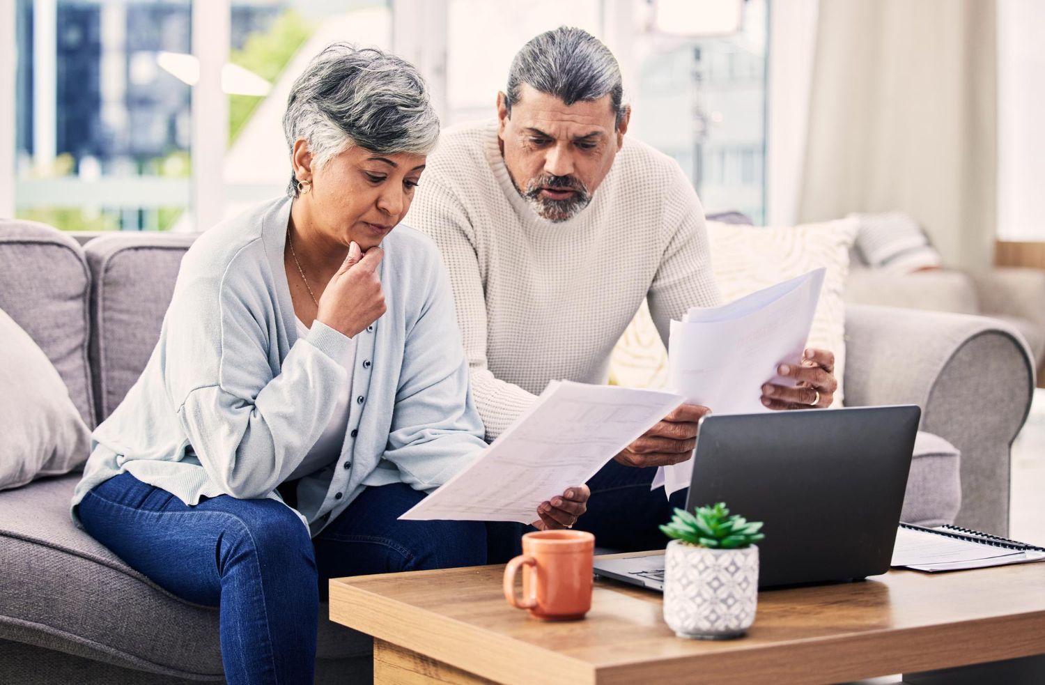 Two people sitting on a sofa looking at documents and a laptop on a coffee table in a brightly lit living room.