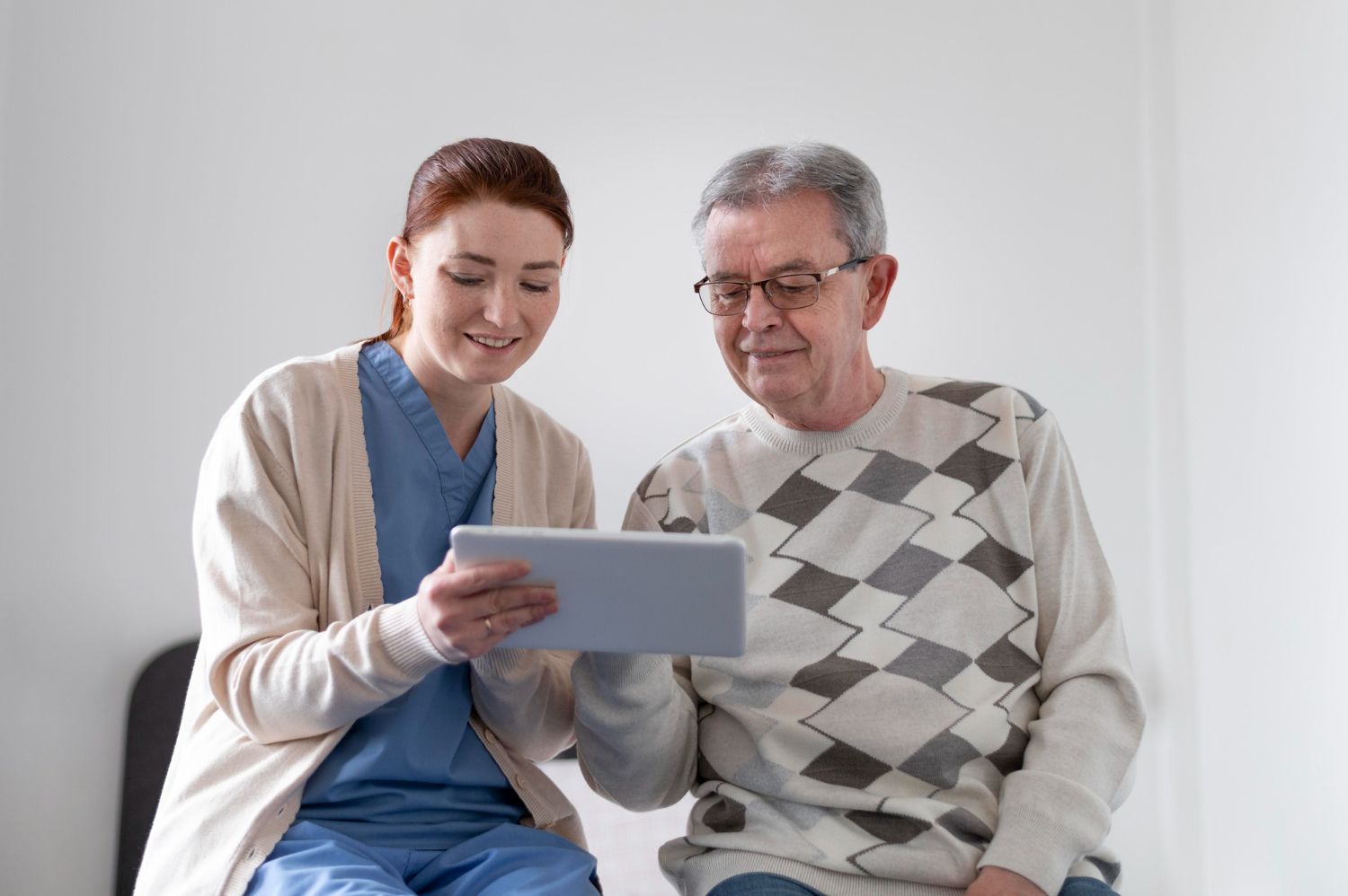 A person in blue scrubs and a cardigan shows a tablet to an older person wearing a patterned sweater.