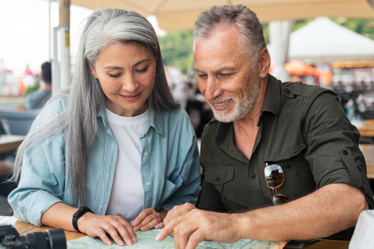 A couple sits at an outdoor table, smiling as they study a paper map together.