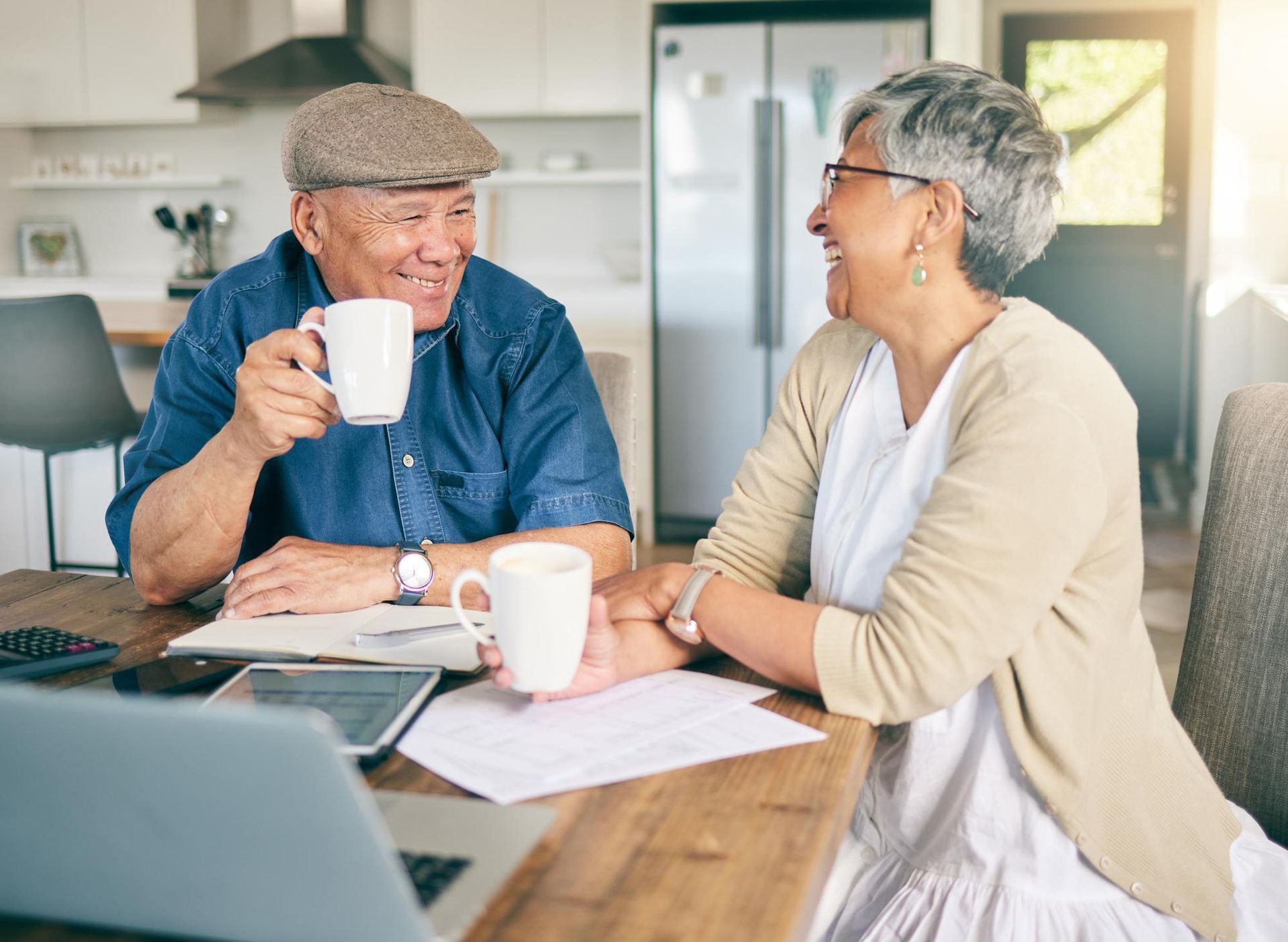 Two people sit at a wooden table in a bright kitchen, smiling at each other while holding mugs of coffee near a laptop.