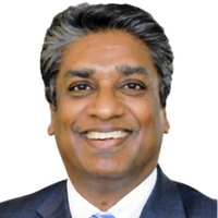 A smiling man with dark hair wears a dark suit and light blue tie against a white background.