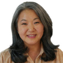 A portrait of a smiling person with shoulder-length, dark hair, wearing a tan shirt against a white background.