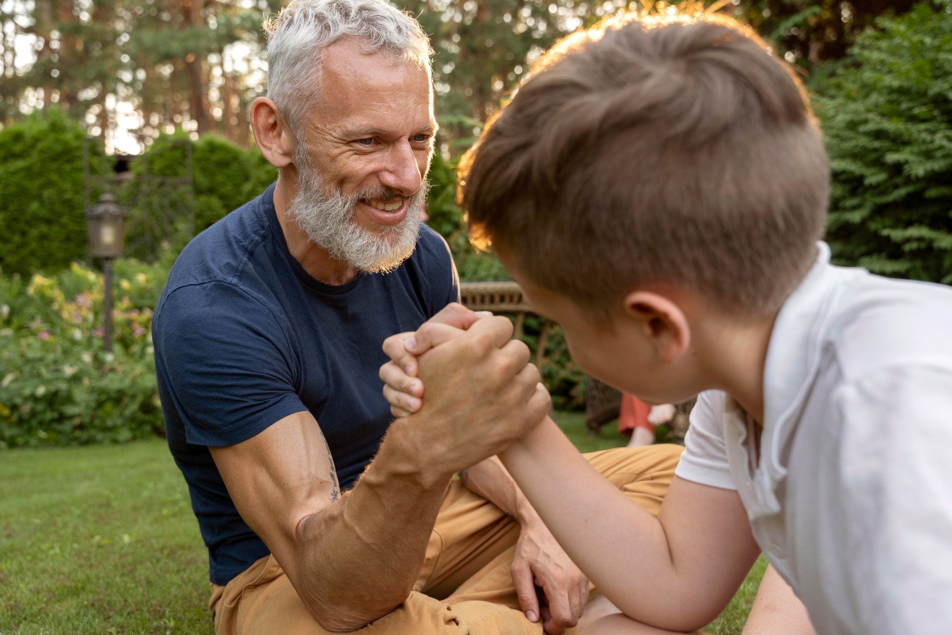 An older adult and a child arm wrestling on a lawn in the sunlight.
