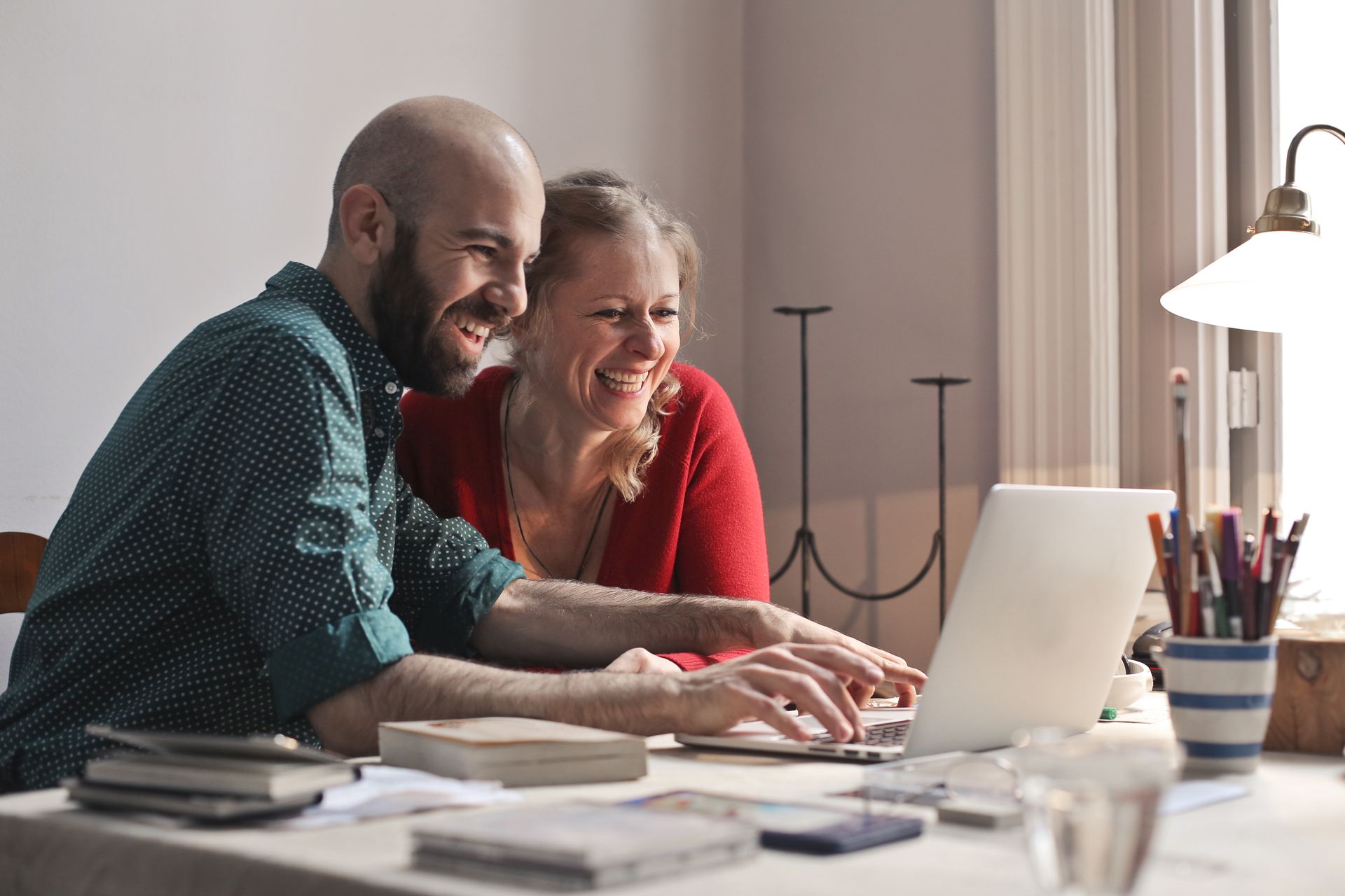 A man and woman sit at a table, smiling while looking at a laptop computer together in a bright, indoor setting.