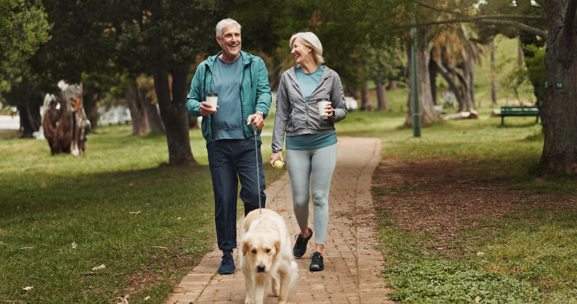 Two people walking a golden retriever on a paved park path, both holding paper cups and smiling at each other.