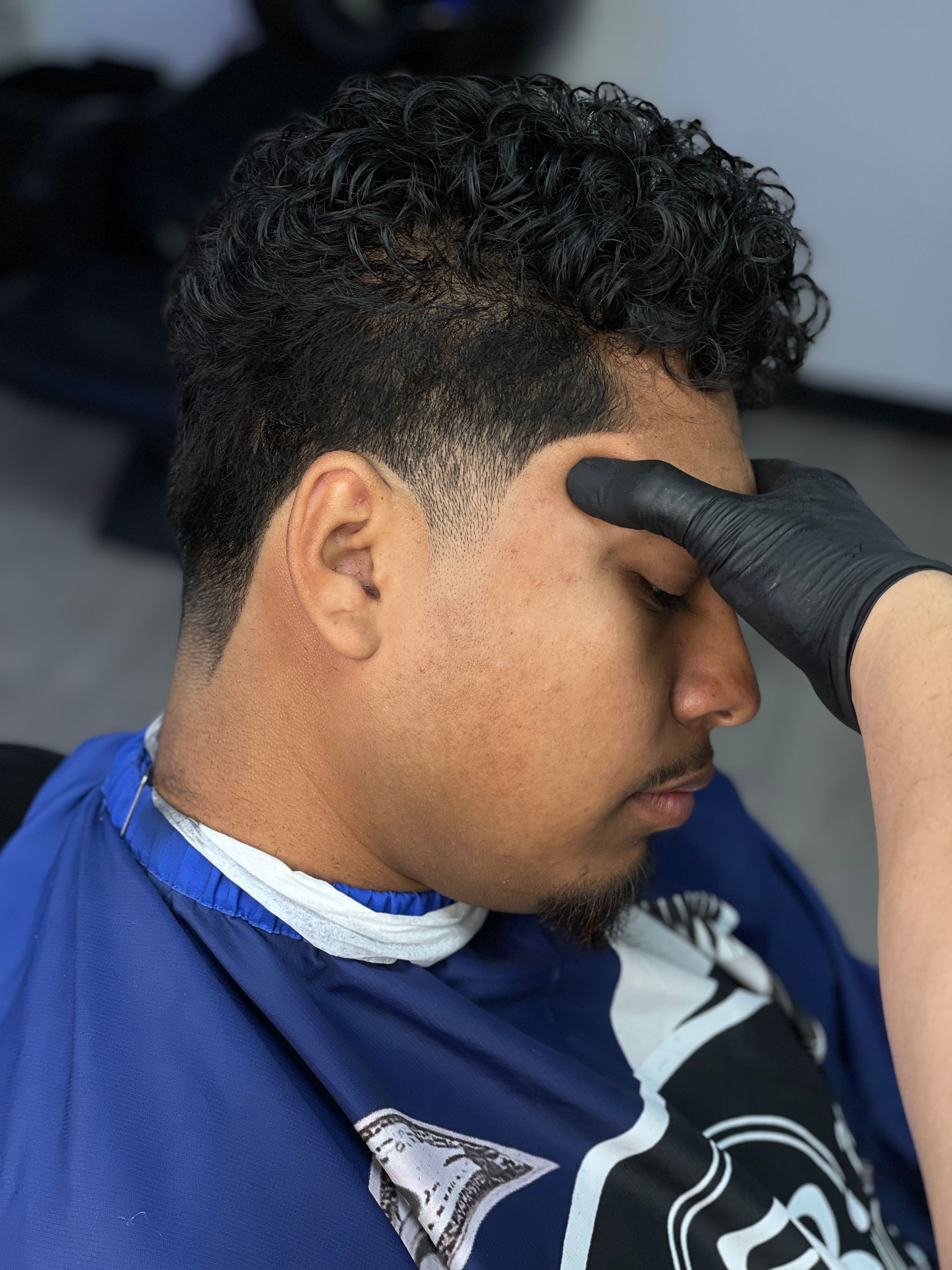 Person with curly hair getting a haircut. Barber's gloved hand on forehead, blue cape, clean fade.