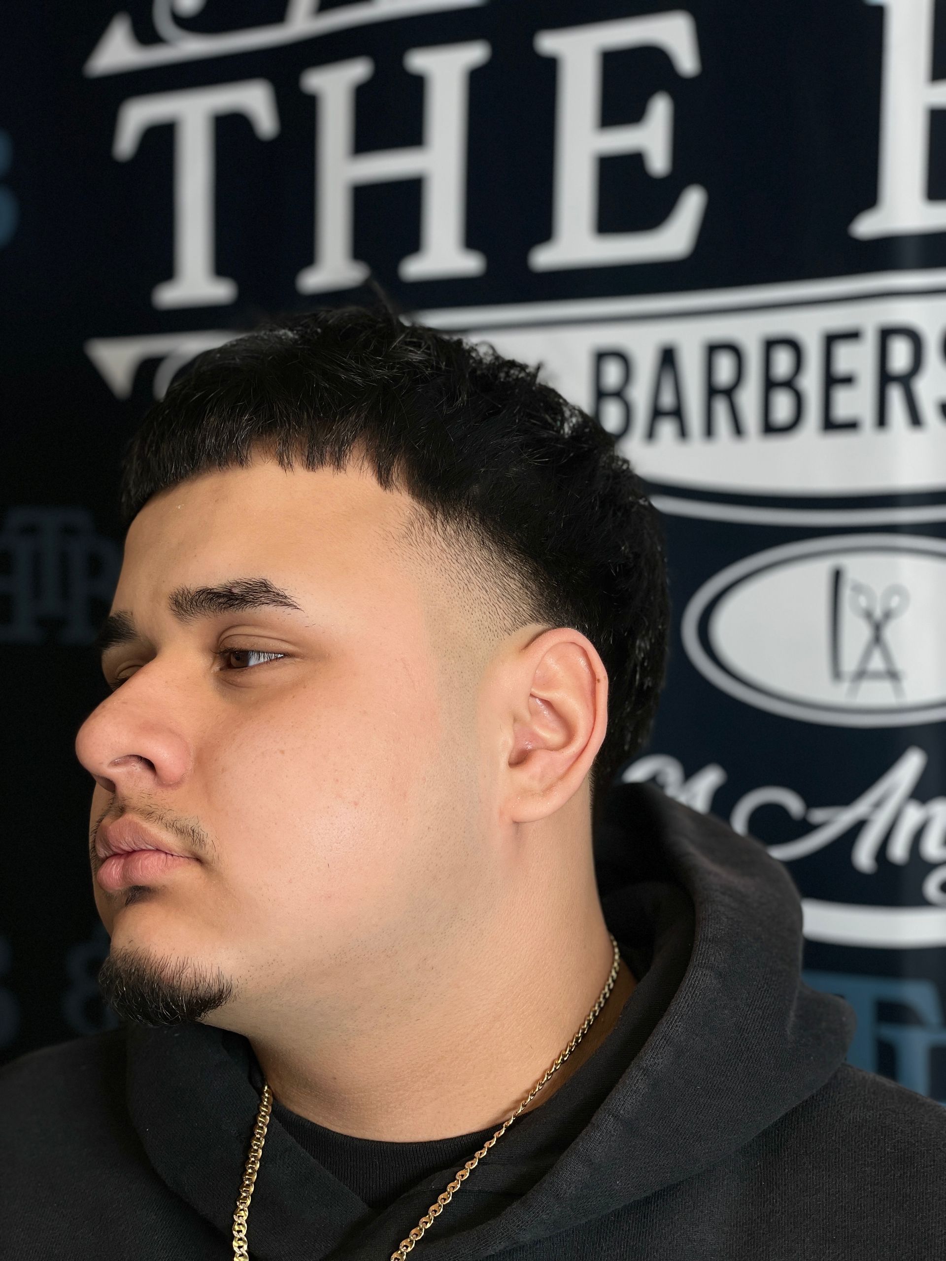 Man with a faded haircut, stubble, and gold chain, in a barbershop setting.