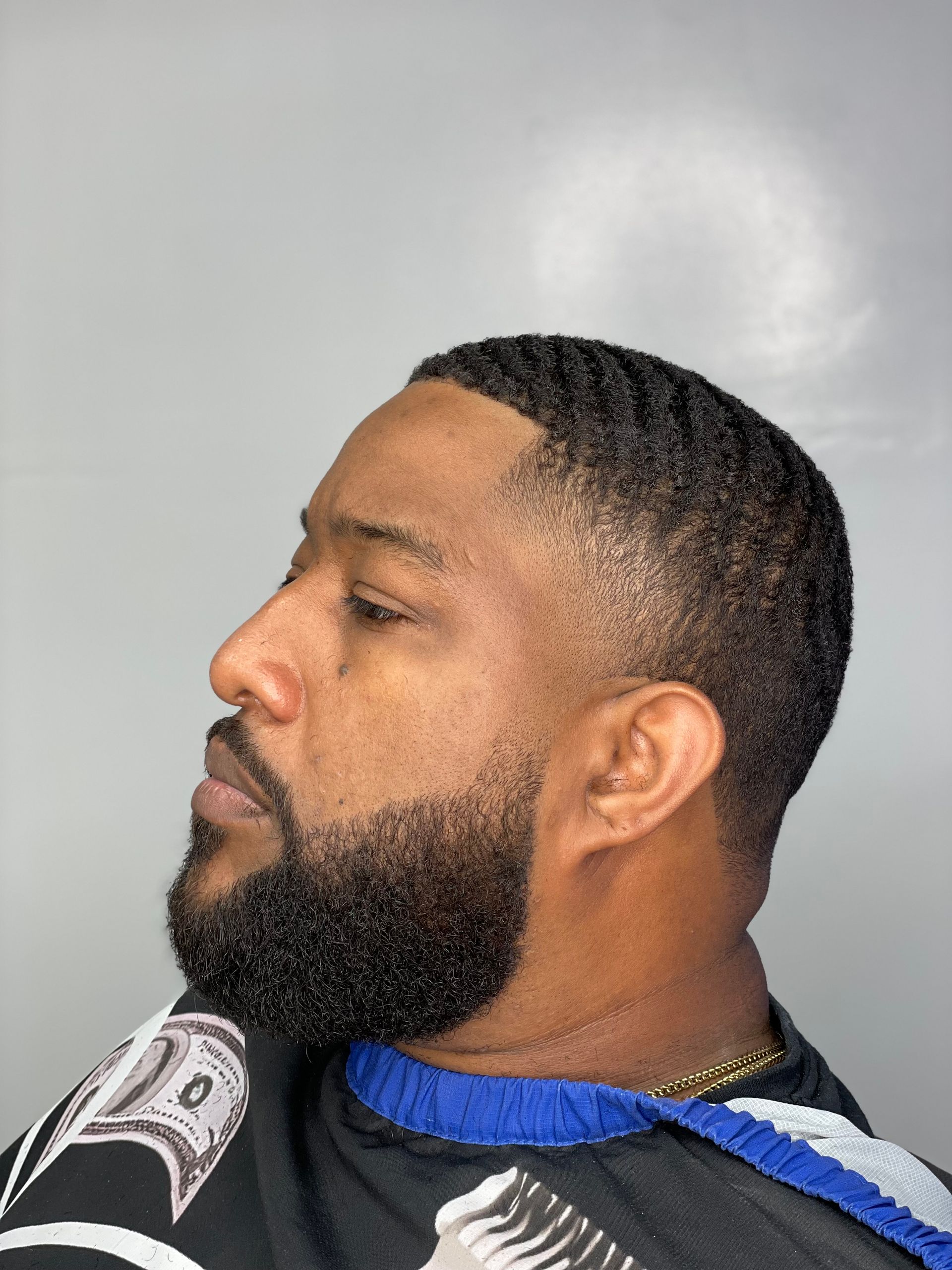 Man with a short haircut and beard, viewed in profile. The hair is wavy and faded. He wears a cape.