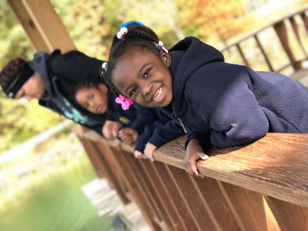A little girl is leaning on a wooden railing and smiling.