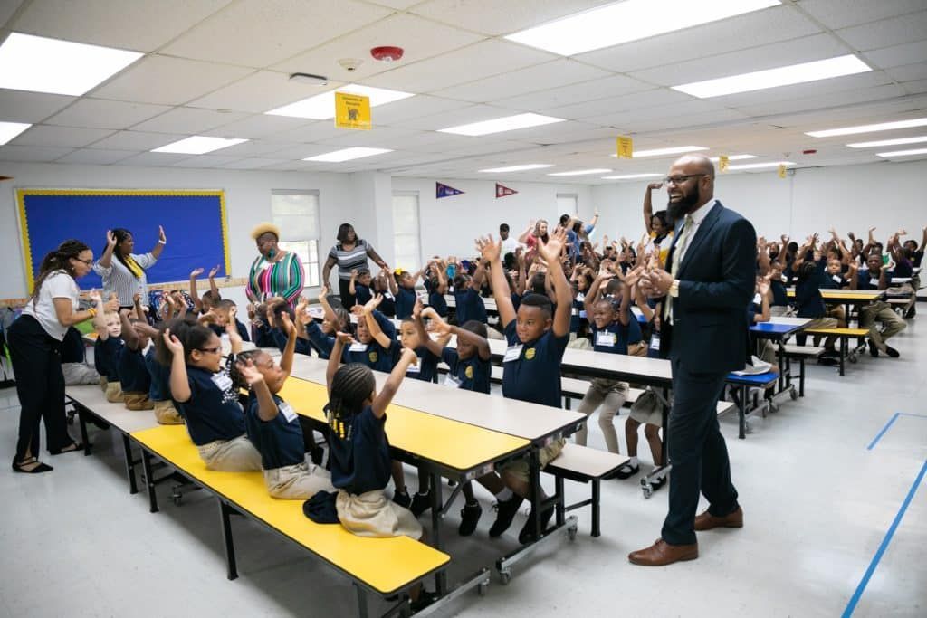 A man is standing in front of a large group of children in a school cafeteria.