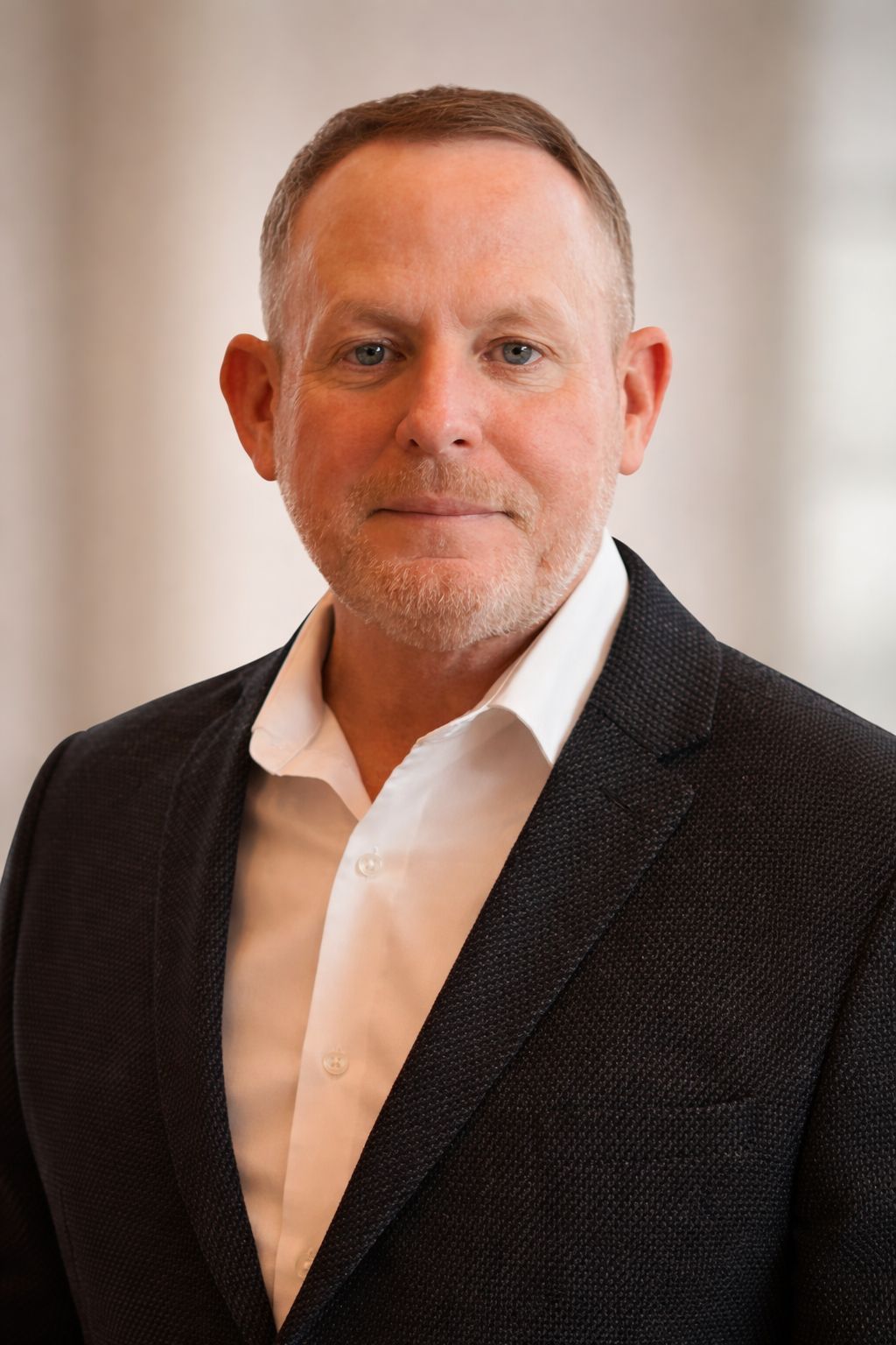 Man in dark blazer and white shirt, looking forward, neutral expression, indoors.