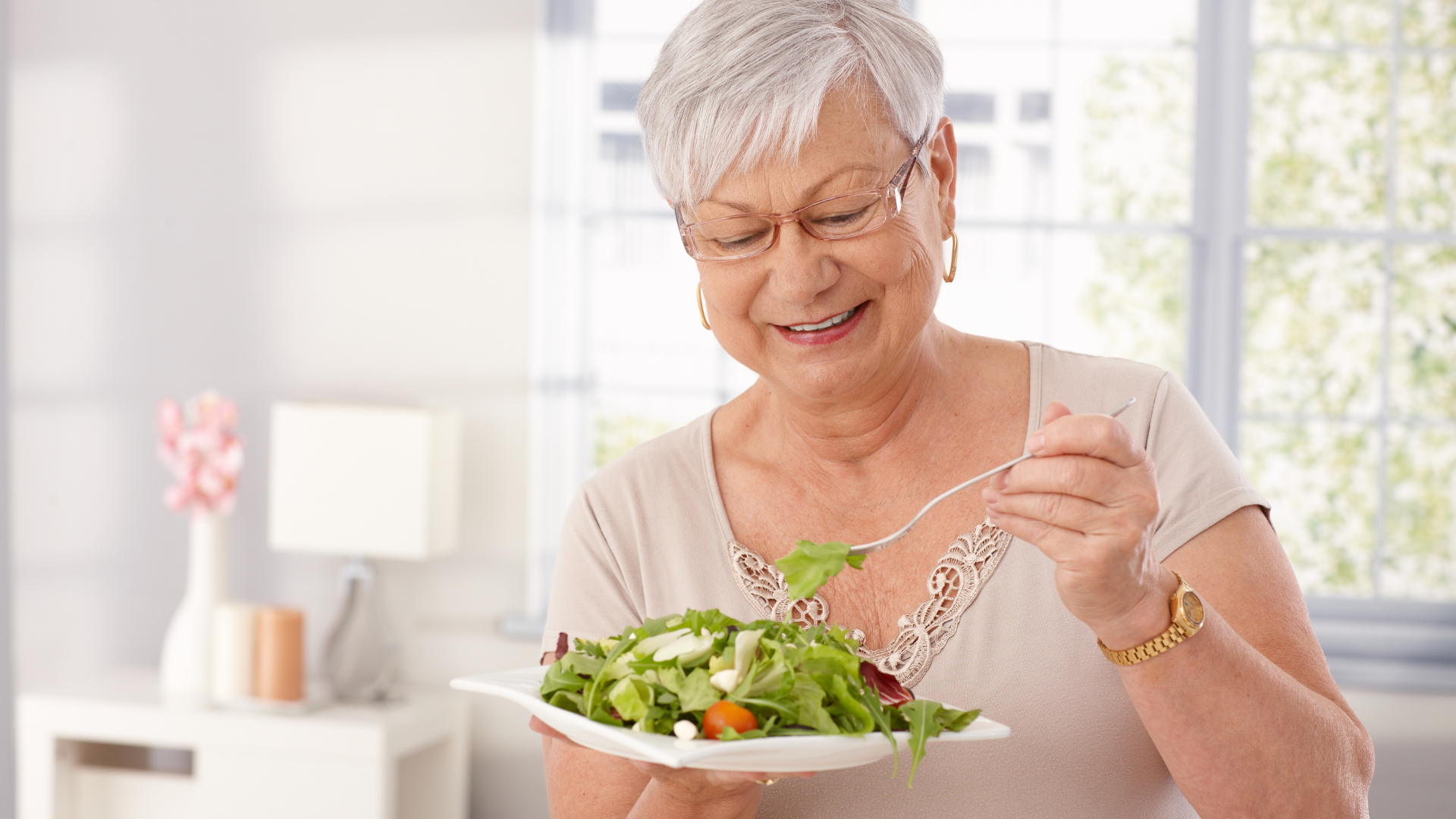 Woman eating a salad from a white plate indoors. She wears glasses and smiles.