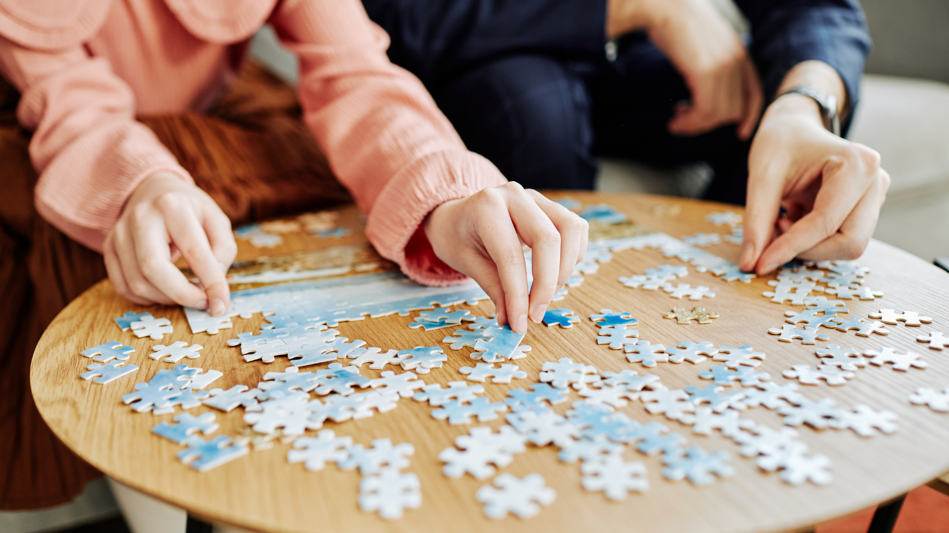 Two people assembling a blue and white jigsaw puzzle on a wooden table.