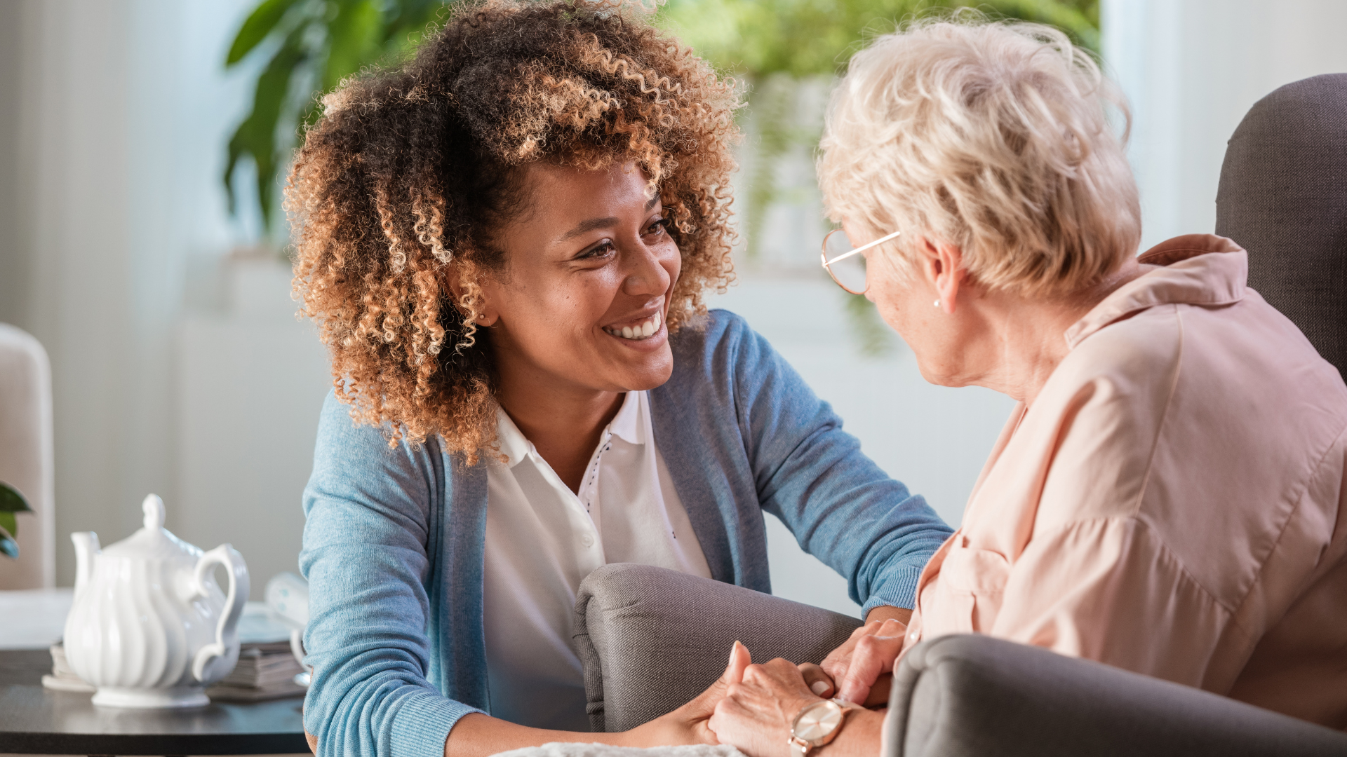 Woman with curly hair smiles, holding hands with an older woman seated indoors.