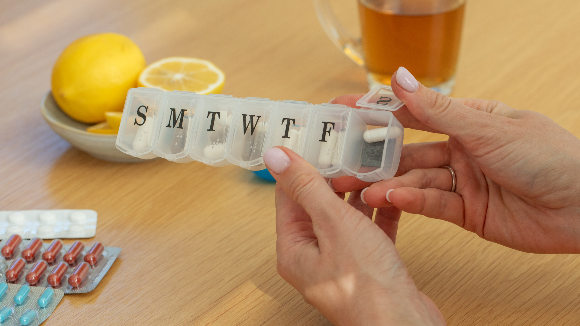 Hands holding a pill organizer. Lemon, tea, and medication packs sit on a wooden table.