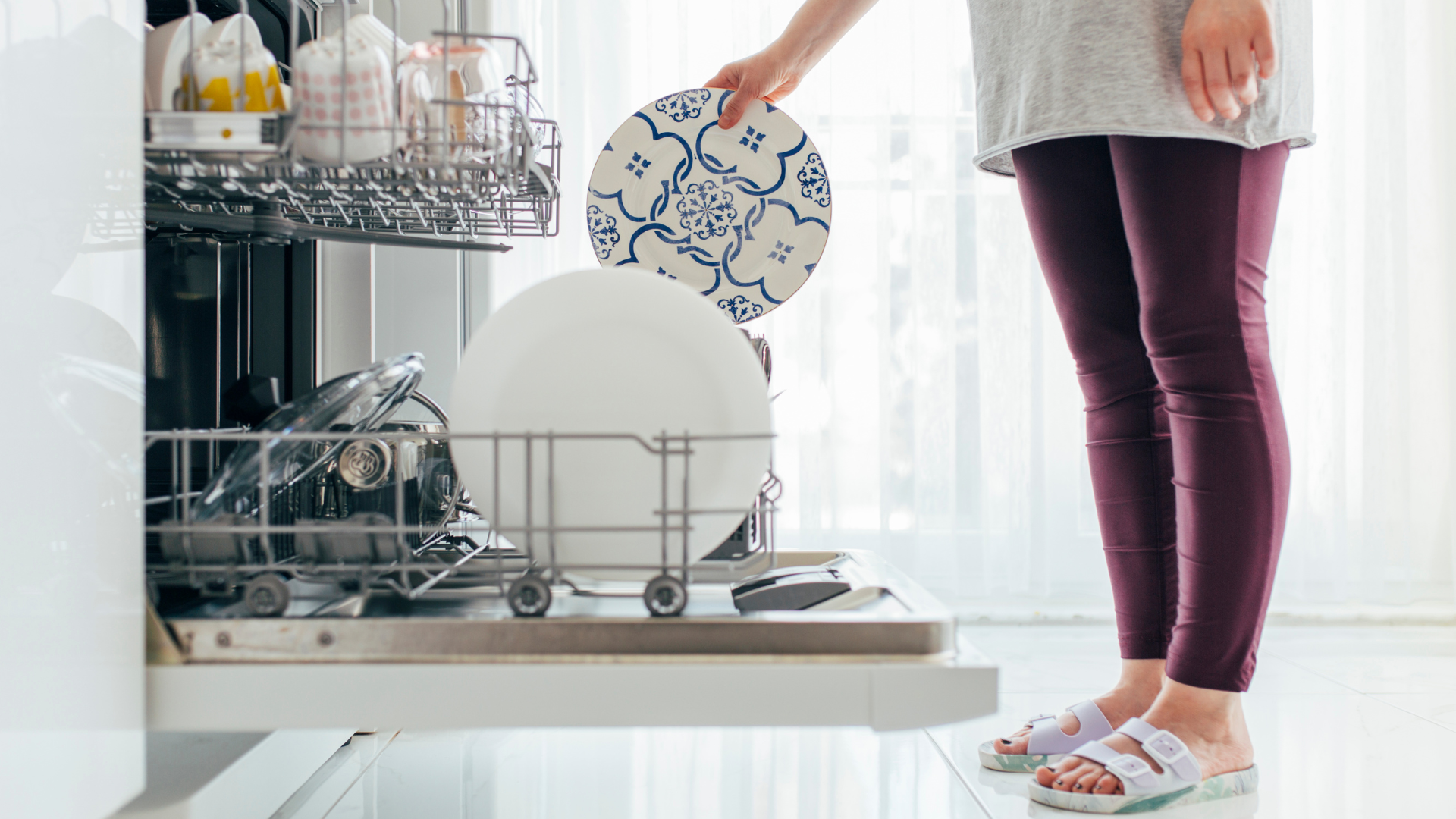 Person loading a dishwasher with a plate in a bright kitchen.