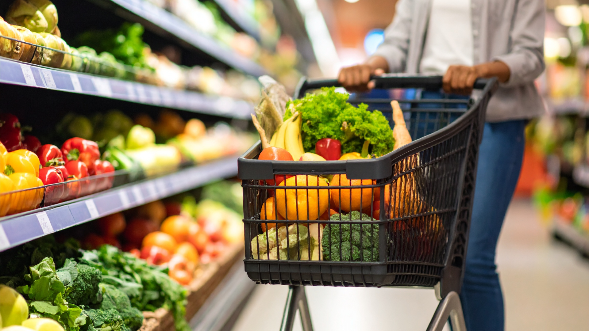 Person pushing a grocery cart filled with fruits and vegetables in a supermarket aisle.
