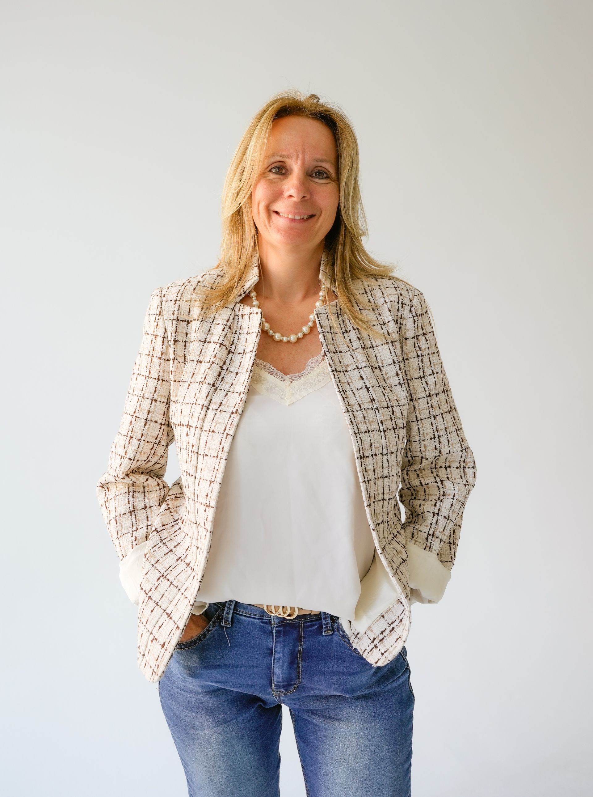 Woman in a blazer and jeans smiles with hands in pockets.