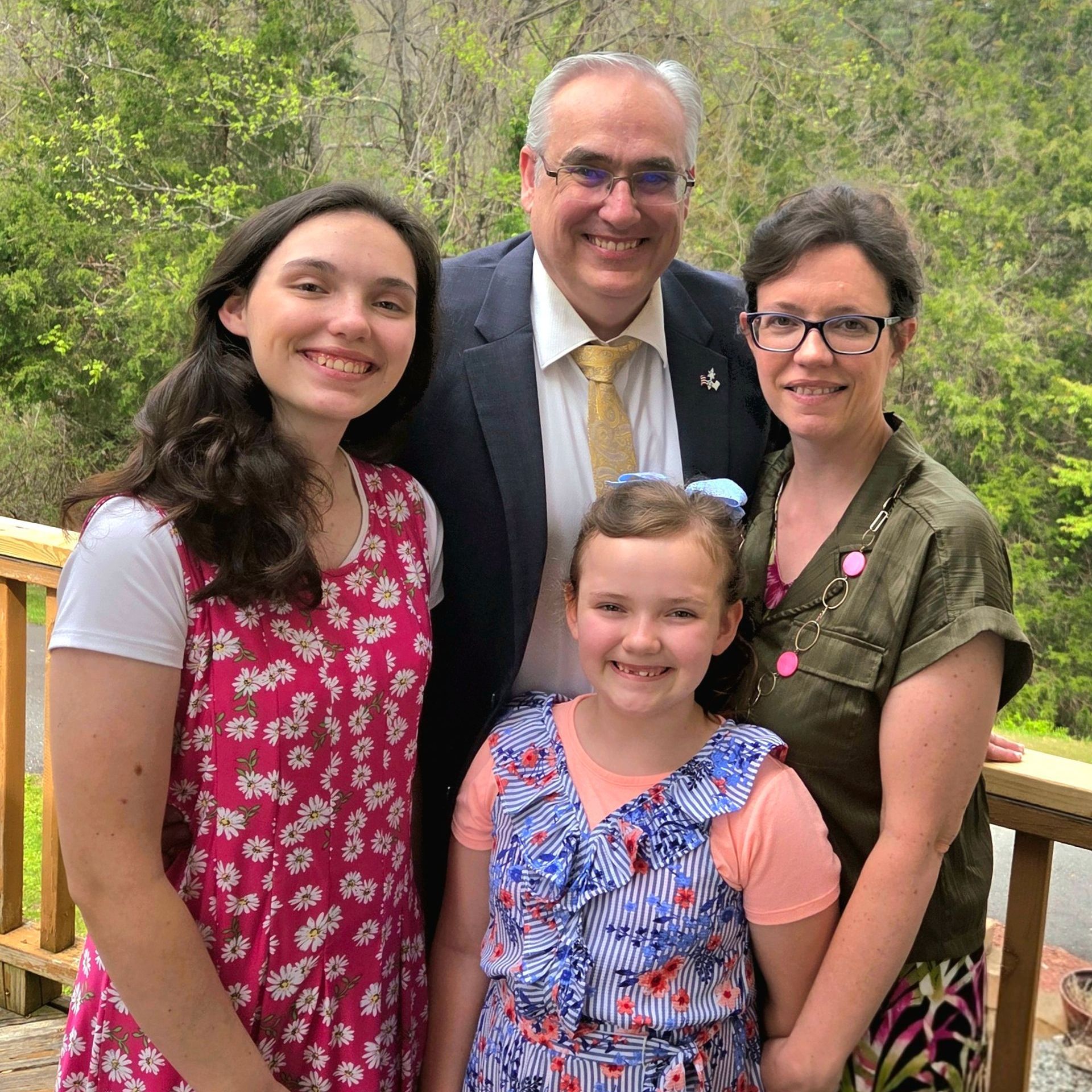 Family of four smiles on a wooden deck. Father in suit, two daughters, mother in glasses. Outdoors.