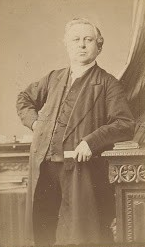 Man in suit, standing by desk, arm on hip. Light hair, serious expression, sepia tone.