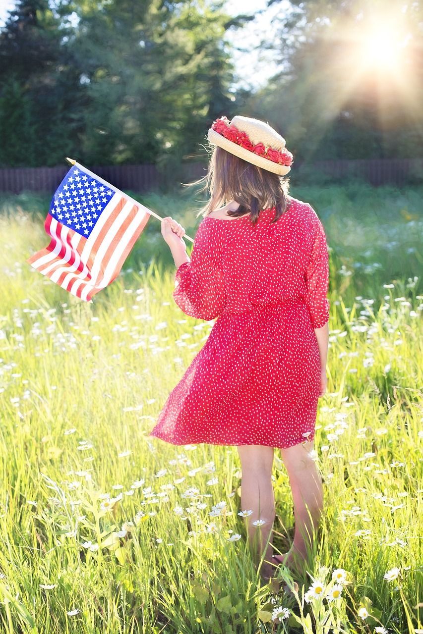 Woman in red dress holding American flag in a sunny field of wildflowers.