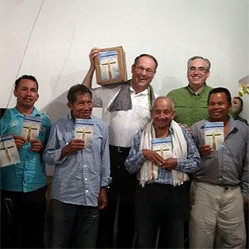 Group of men smiling and holding books with a cross. Indoors, neutral backdrop.