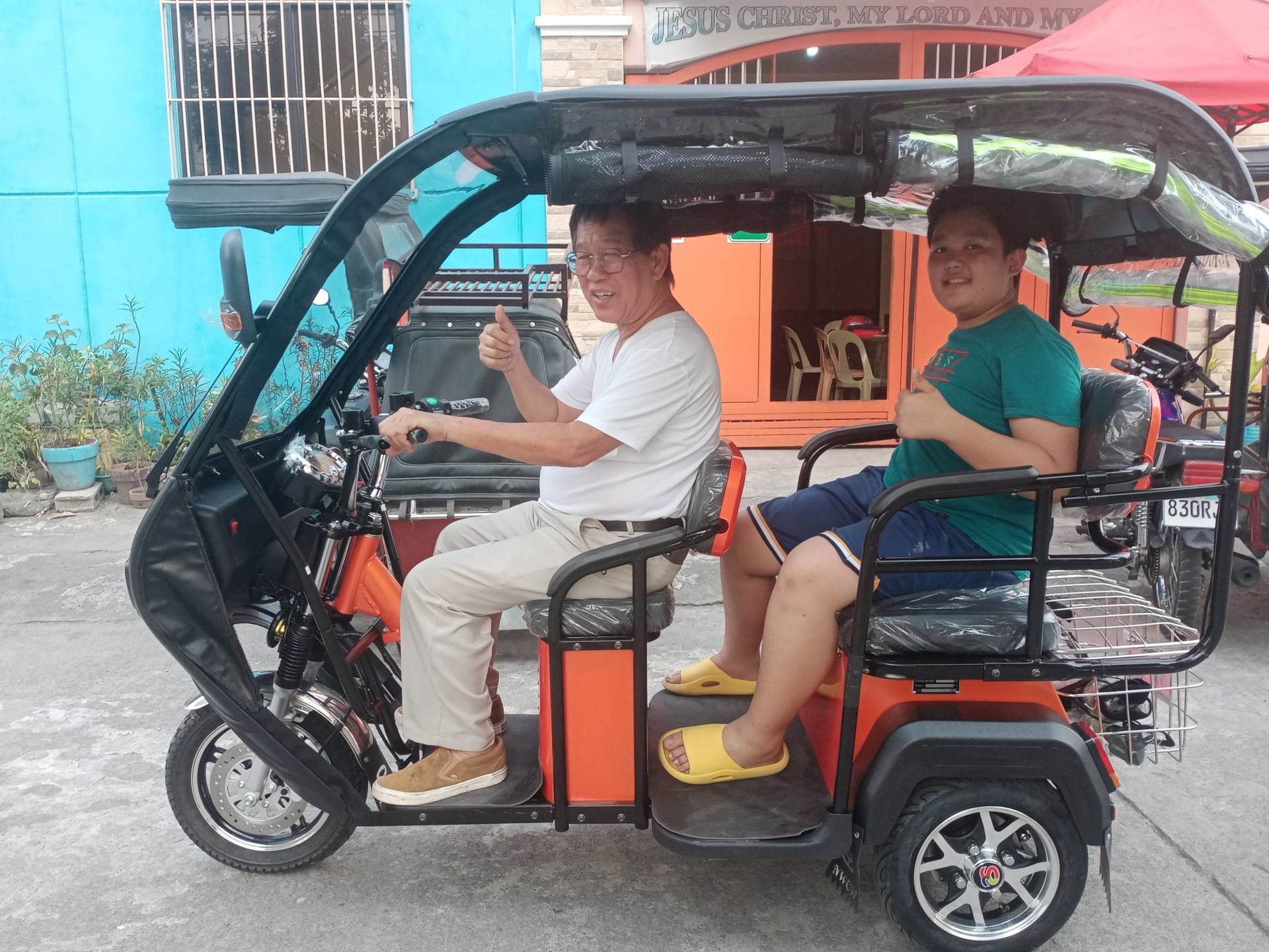 Two men in an orange electric tricycle, one driving, the other as a passenger, both giving thumbs up.