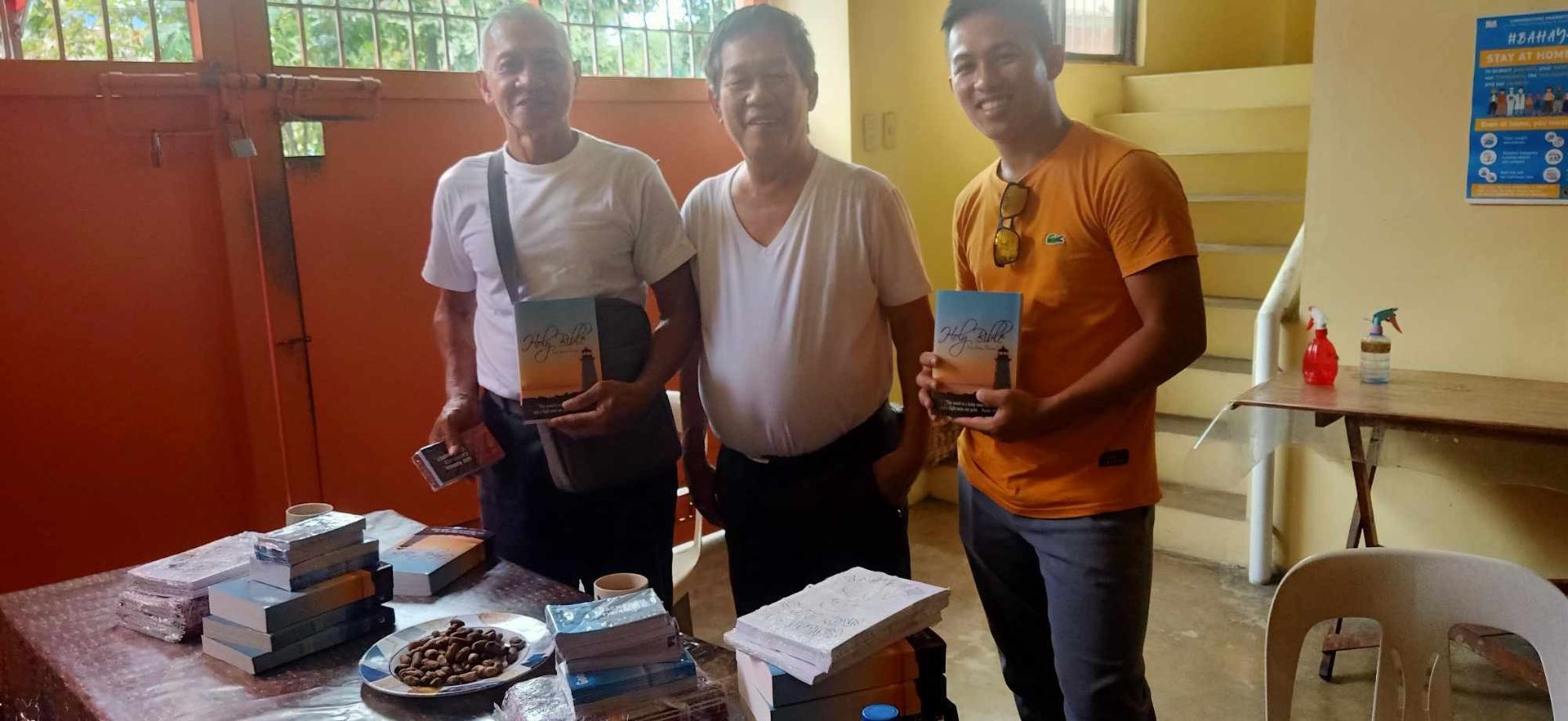 Three men holding books at a table with stacks of books. They are inside a room with orange walls.