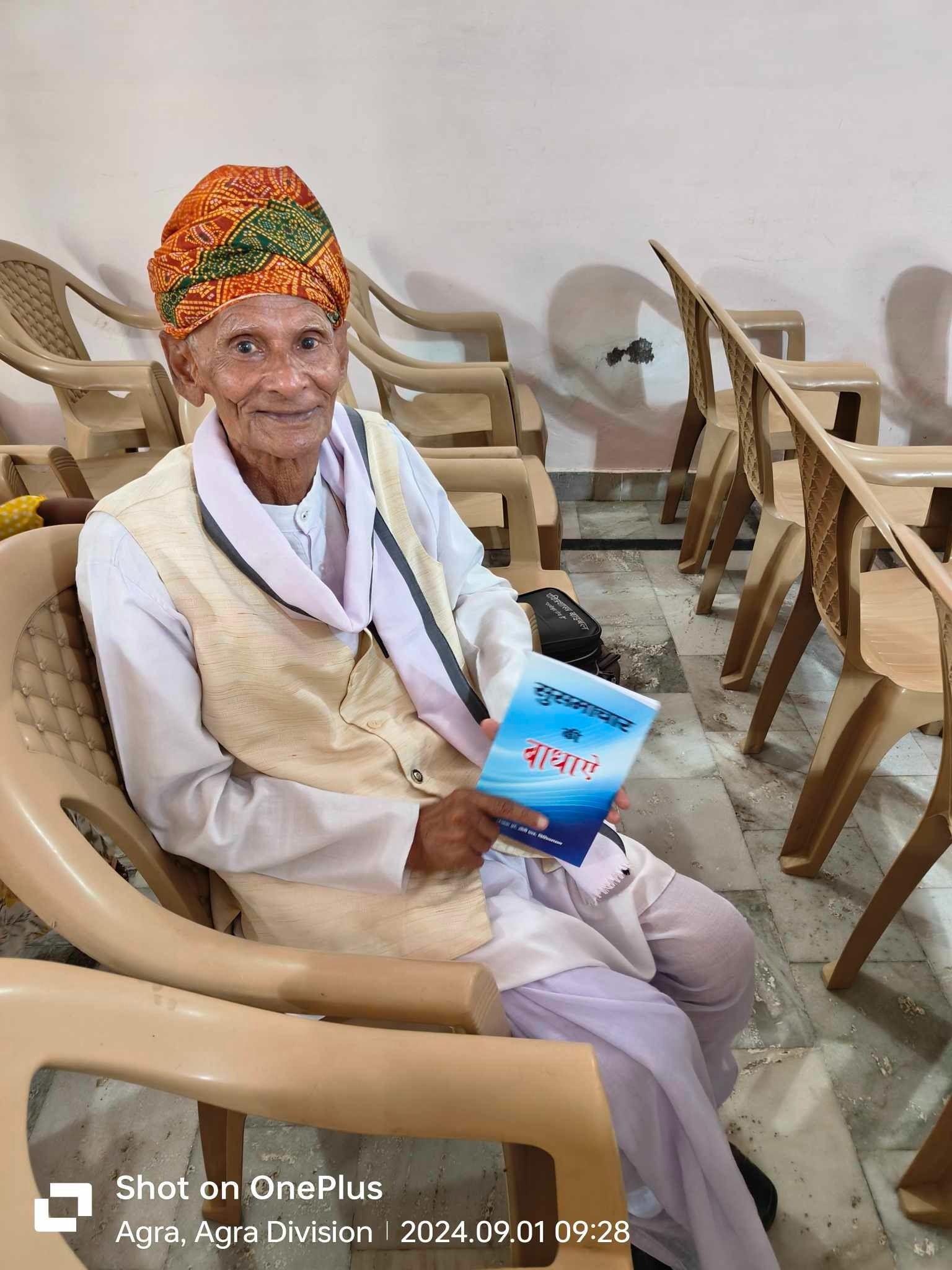 Elderly man in traditional wear, holding a book, seated in a room with chairs.