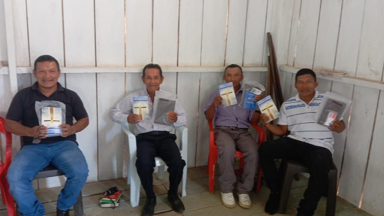 Four men seated, each holding a book with a cross on it. They're indoors.