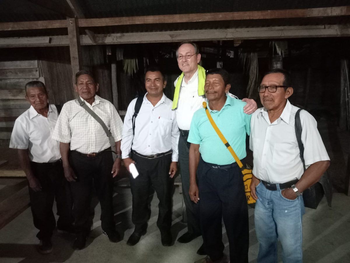 Group of six men smiling, posing inside a wooden structure, possibly a community meeting.