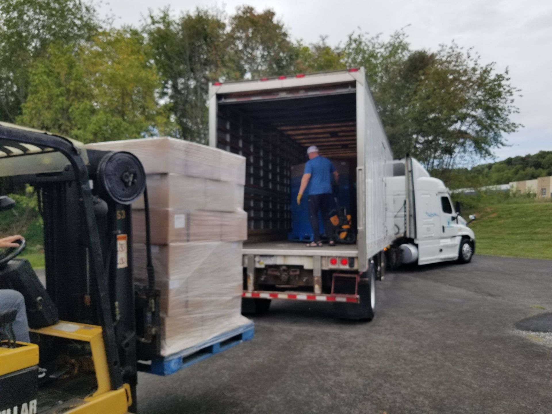 Forklift loading boxes onto a semi-truck trailer; a person inside the trailer is organizing the cargo; outdoors.