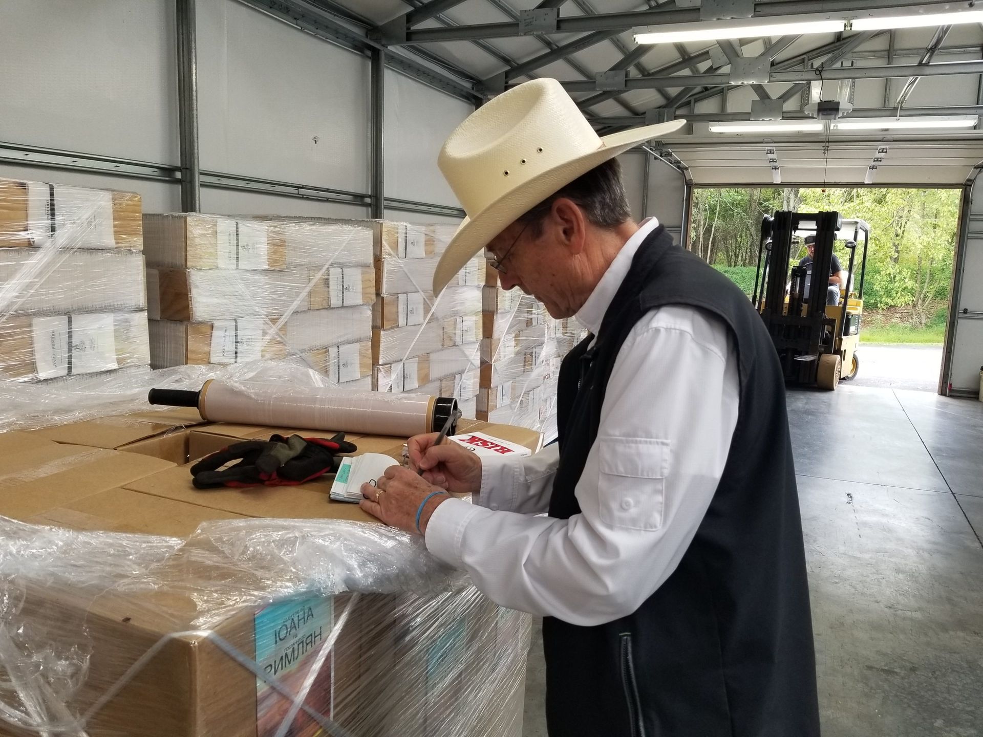 Man in cowboy hat signing paperwork on a wrapped pallet in a warehouse with a forklift in the background.