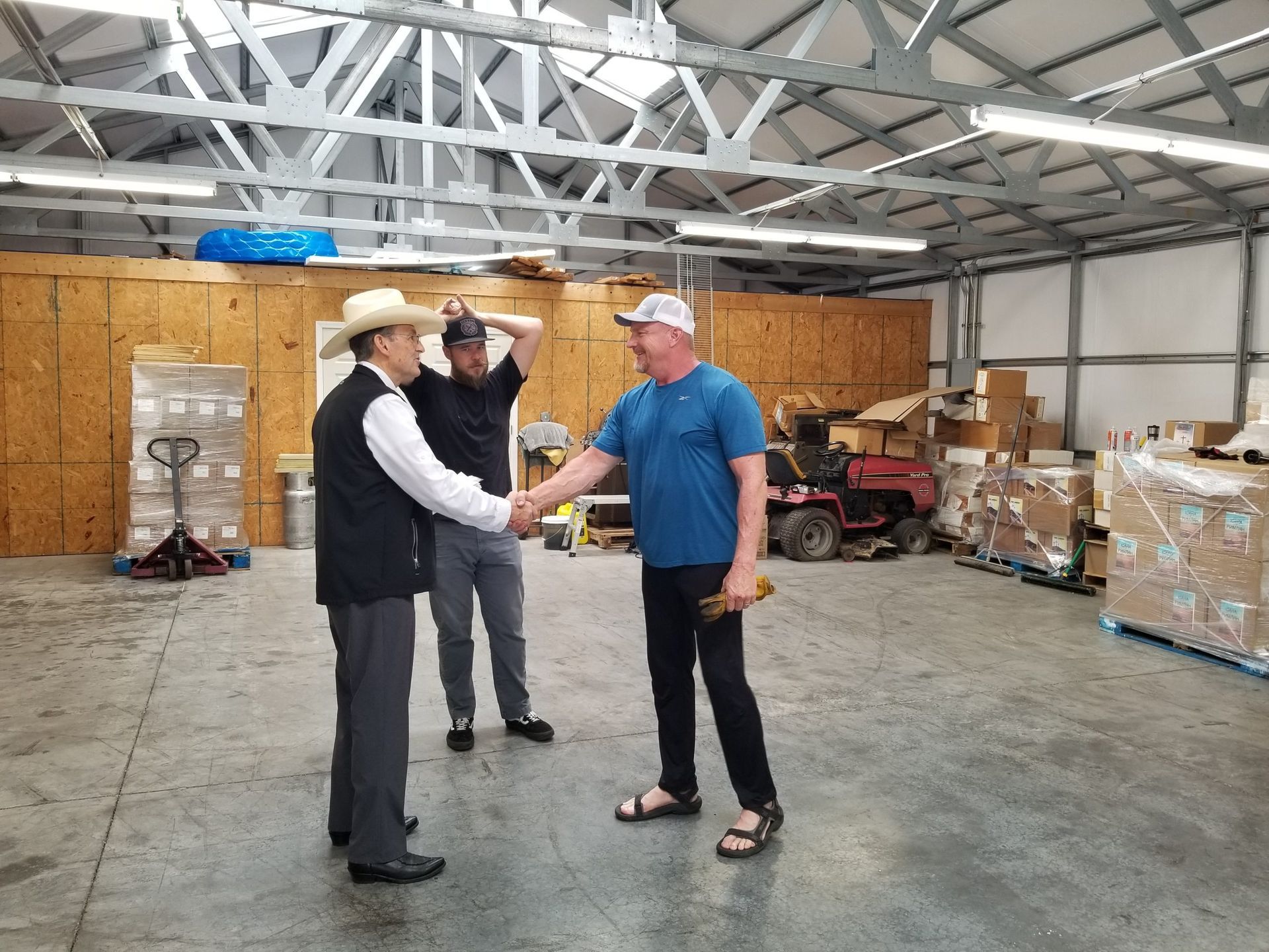 Three men shake hands in a warehouse; one wears a hat and vest. Boxes and machinery are visible.