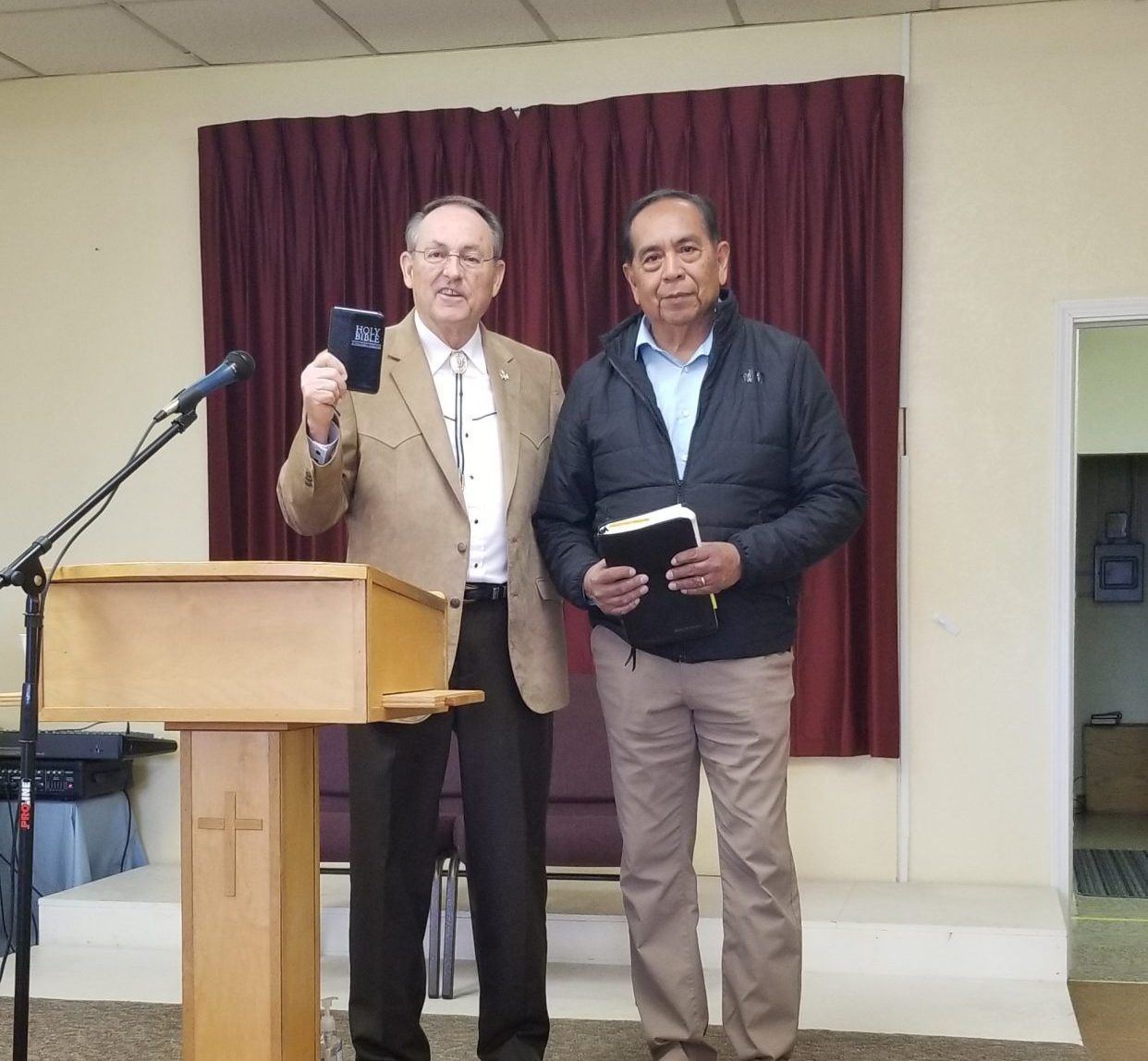 Two men standing next to a podium in front of a burgundy curtain, each holding a bible.