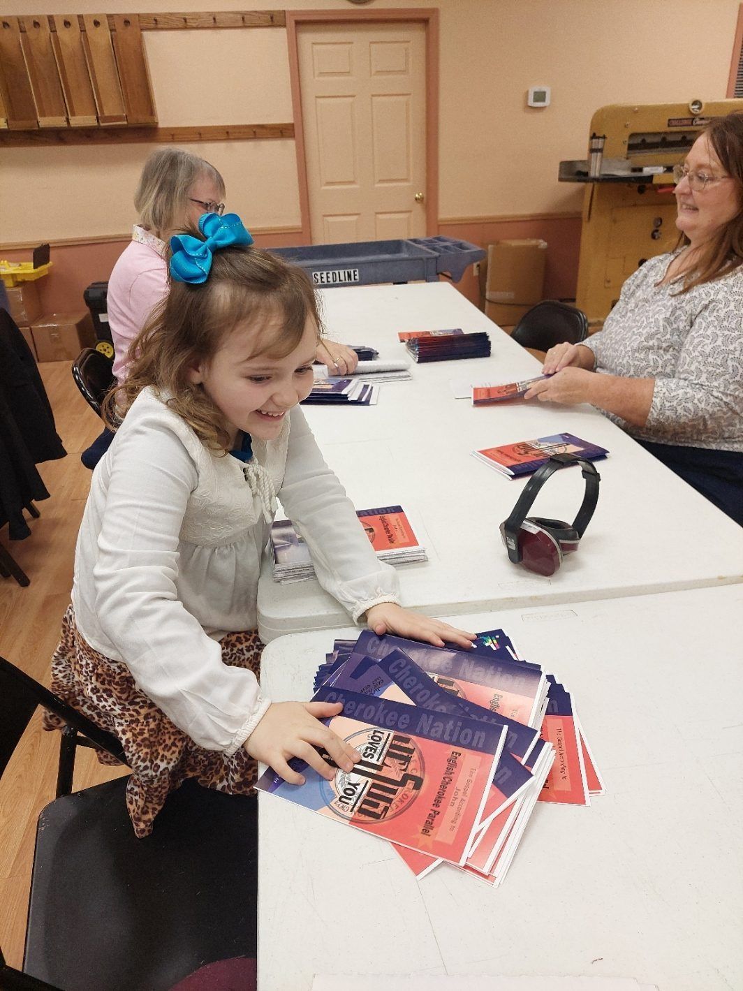Young girl smiles while holding flyers at a table; two women are seated beside her.