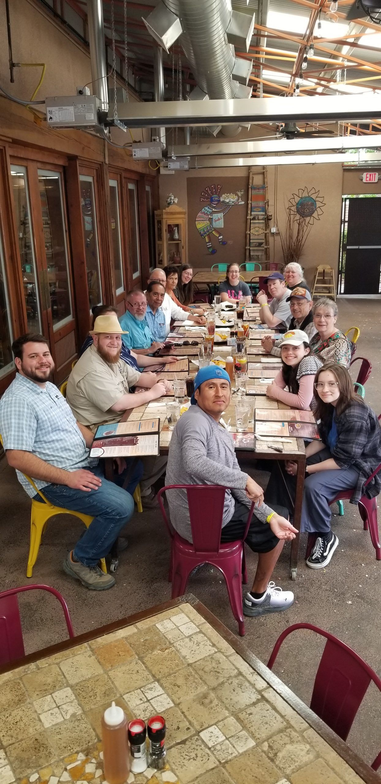 People sitting at a long table in an outdoor dining area. Some are facing the camera. The table is set for a meal.