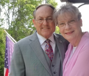 Smiling man and woman, likely a couple, posing outdoors. Man in suit, woman in pink. American flag visible.