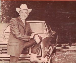 Man in cowboy hat leans on a vintage car, smiling. Wearing a suit and boots.