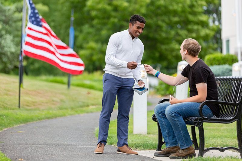 Two men, one standing and handing something to the seated man on a bench near an American flag.