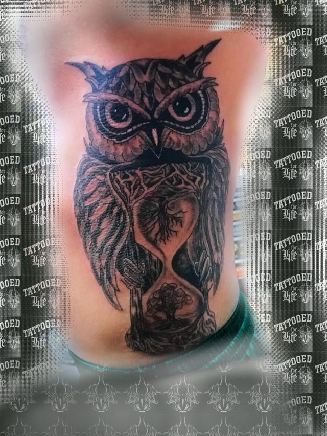 A black and white tattoo of an owl and an hourglass on a person 's back.