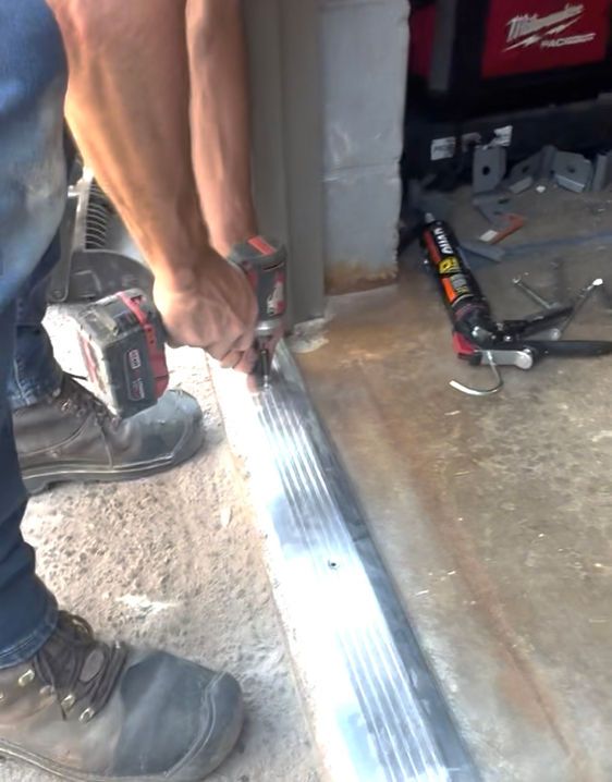 Person using a drill to install a garage door threshold.  Tool, sealant tube, and concrete floor visible.