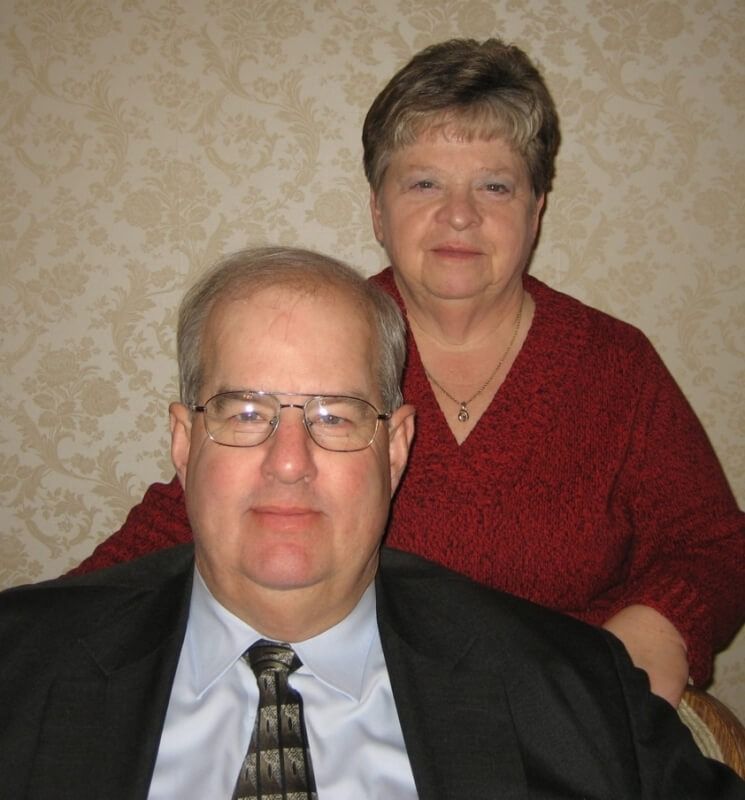 Man in suit and woman in red sweater posing together; indoor setting.