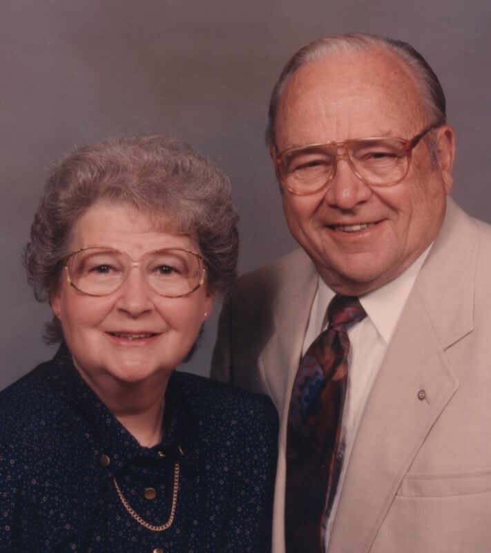 Elderly couple, woman in blue, man in beige suit, both wearing glasses, smiling.