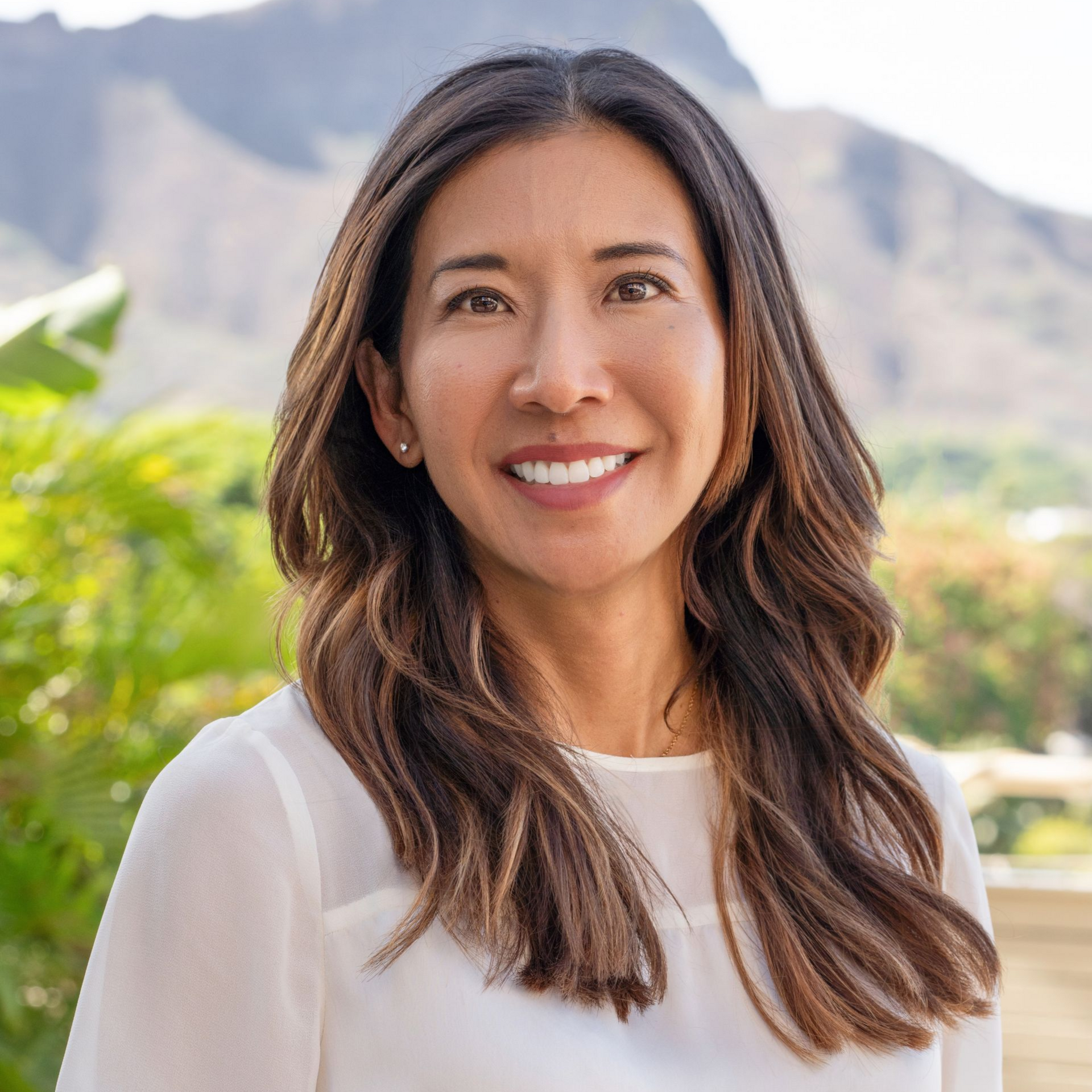 Woman with long brown hair smiling outdoors with a mountain backdrop.