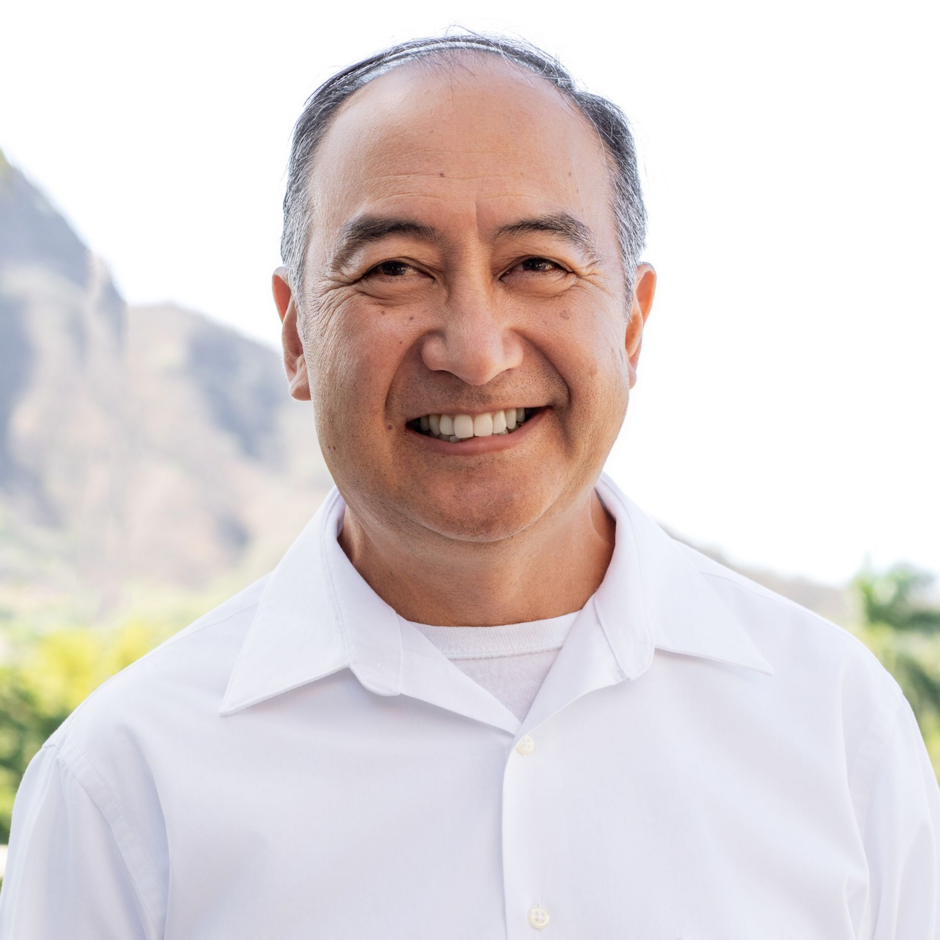 Man with dark hair, wearing a white shirt, smiling. Mountain in background.