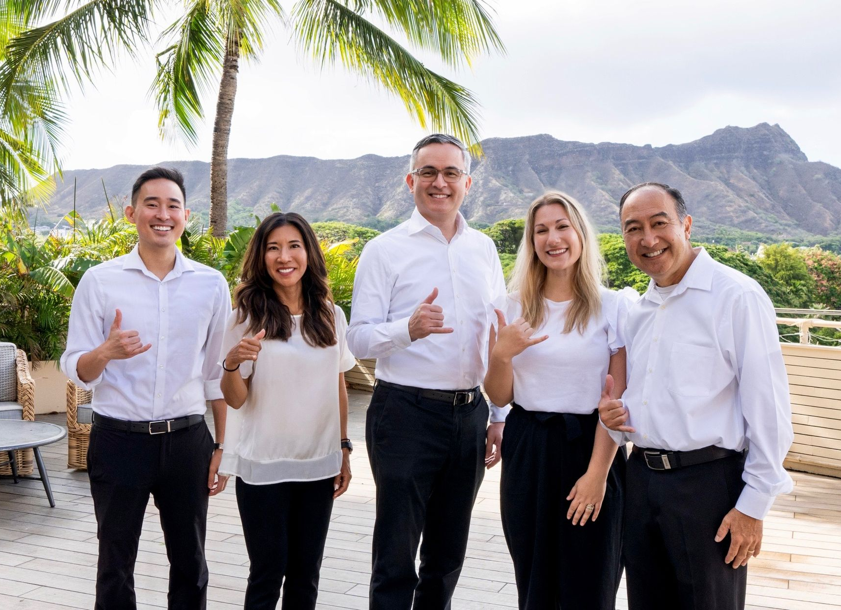 Five people in white shirts and dark pants pose outside with Diamond Head in the background, making shaka signs.