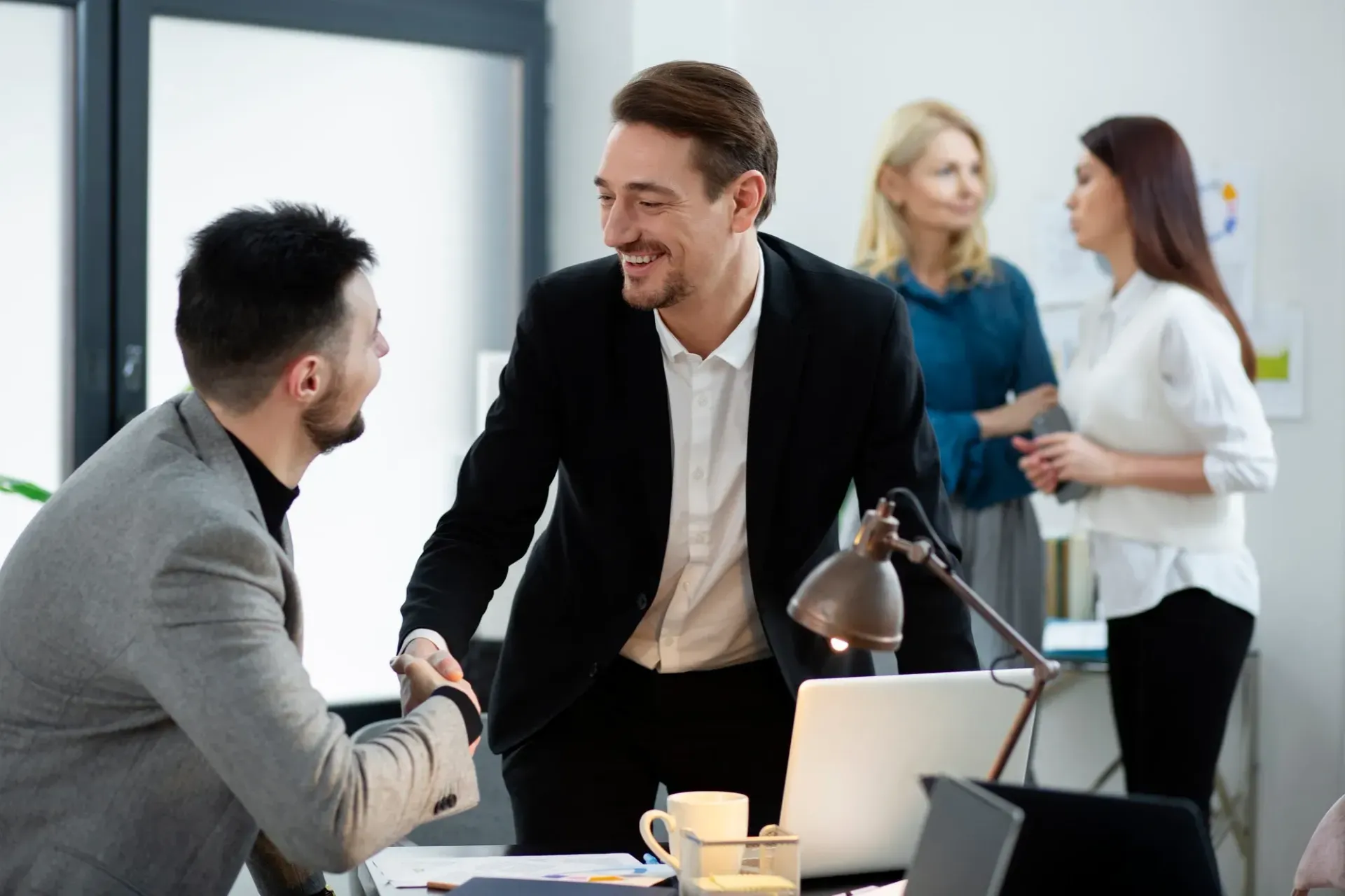 Two men shaking hands in office; two other people conversing in the background.